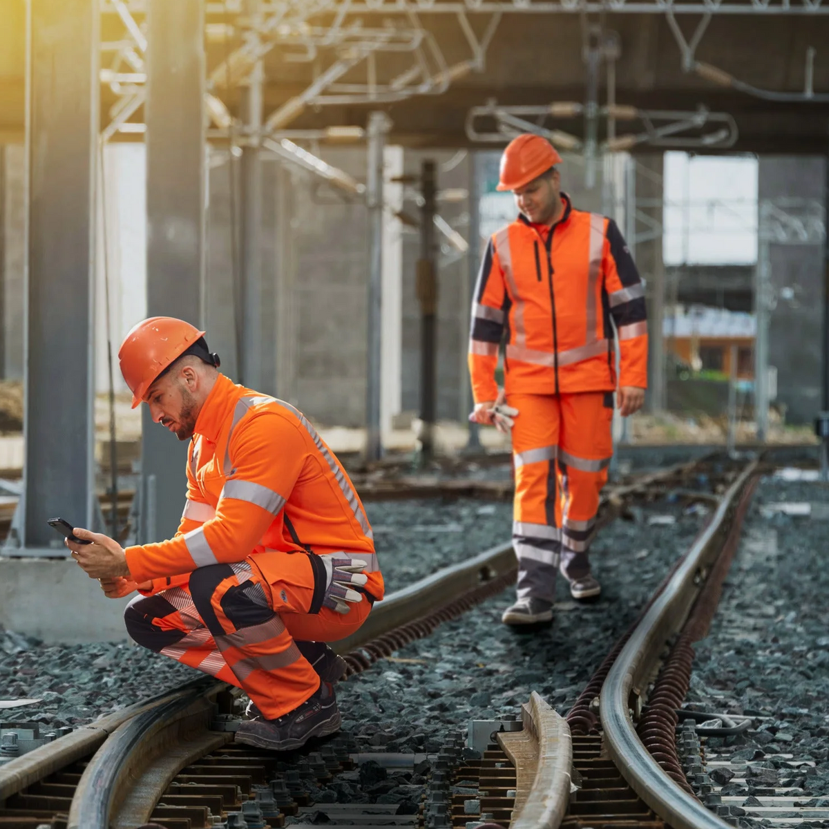 Zwei Arbeiter in orangefarbener Warnschutzkleidung und Schutzhelmen kontrollieren Gleise auf einer Bahnbaustelle