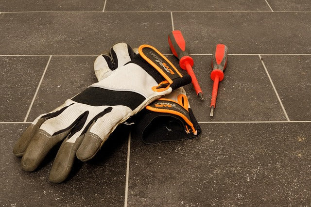 Schutzhandschuhe mit grauen Fingerspitzen neben zwei Schraubendrehern auf einem Fliesenboden