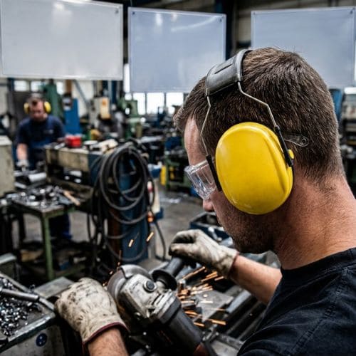 Mitarbeiter mit gelbem Kapselgehörschutz und Schutzbrille arbeitet mit einem Winkelschleifer in einer Werkhalle